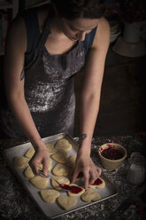 Backen zum Valentinstag, junge Frau steht in einer Küche und verteilt Himbeermarmelade auf herzförmigen Keksen. - MINF06165