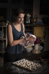 Valentine's Day baking, young woman standing in a kitchen, tray of heart shaped biscuits, wiping her hands with a tea towel. - MINF06170