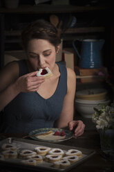 Backen zum Valentinstag, junge Frau sitzt in einer Küche und isst einen herzförmigen Keks. - MINF06172