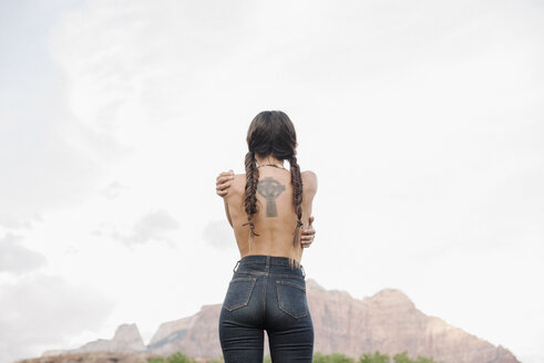 Rear view of a topless young woman with long brown hair in braids, wearing jeans, tattoo of a Celtic Cross on her back. - MINF06280