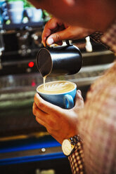 Specialist coffee shop. A barista pouring hot milk in to the froth on a cup of coffee to make a pattern. - MINF06309