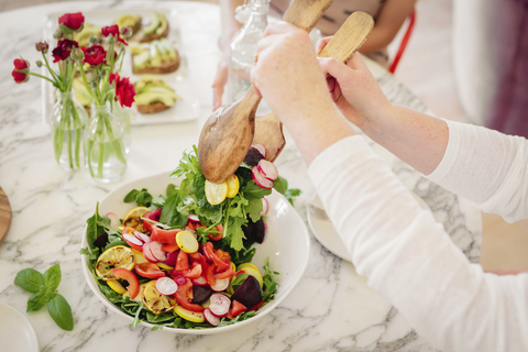 Blick von oben auf einen mit Besteck und Tellern gedeckten Tisch mit vorbereiteten Speisen. Eine Frau nimmt eine Portion Salat., lizenzfreies Stockfoto