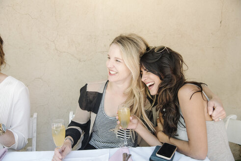 Zwei Frauen, die an einem Tisch sitzen, sich umarmen und lachen, in der Hand Gläser mit Champagner. - MINF06346