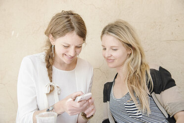 Zwei lächelnde Frauen mit langen blonden Haaren sitzen an einem Tisch und schauen auf ein Mobiltelefon. - MINF06348