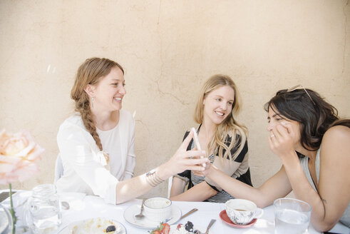 Drei lächelnde junge Frauen sitzen an einem Tisch mit Essen und Getränken und schauen auf ein Handy. - MINF06349