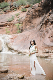 Junge Frau mit langen braunen Haaren, in einem langen weißen Kleid, steht an einem Fluss und hält Arum Lilies. - MINF06350