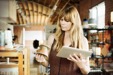 Frau in einem Geschäft, die ein digitales Tablet und einen kleinen Keramiktopf hält. - MINF06363