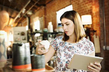 Junge Frau in einem Geschäft, in der Hand ein digitales Tablet und eine Keramiktasse. - MINF06369