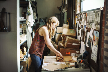 Frau mit langem blondem Haar steht im Lagerraum eines Geschäfts und wickelt Waren in braunes Papier ein. - MINF06399