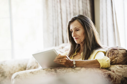 Ältere Frau mit langen braunen Haaren sitzt auf einem Sofa und benutzt ein digitales Tablet. - MINF06431