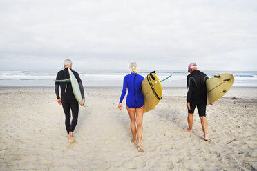 Ältere Frau und zwei ältere Männer an einem Strand, in Neoprenanzügen und mit Surfbrettern. - MINF06460