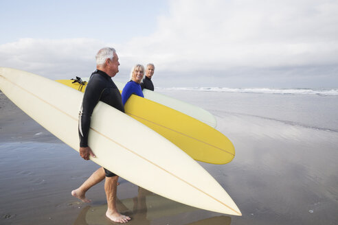 Ältere Frau und zwei ältere Männer an einem Strand, in Neoprenanzügen und mit Surfbrettern. - MINF06461