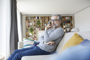 Nachdenklicher reifer Mann sitzt auf der Couch in seinem Wohnzimmer und schaut aus dem Fenster - RBF06486