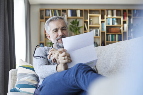 Mature man sitting on couch at home reading letter - RBF06490