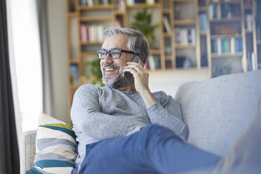 Mature man on the phone sitting on couch at home looking out of window - RBF06491
