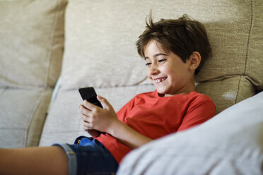 Portrait of laughing little girl lying on the couch looking at cell phone - JSMF00387