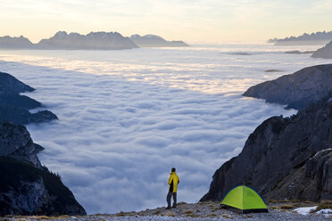 Mann steht neben einem Zelt, das über den Wolken auf einem Berggipfel aufgebaut ist. - MINF06558