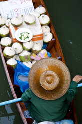 High angle view of person wearing hat sitting in narrow boat, transporting green coconuts. - MINF06615