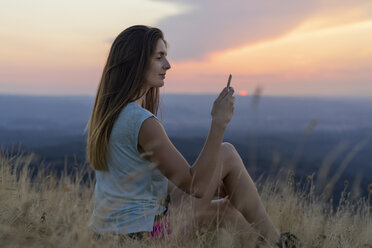 Spanien, Katalonien, Montcau, junge Frau mit Handy sitzt im Gras auf der Spitze des Hügels bei Sonnenuntergang - AFVF01362