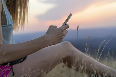 Nahaufnahme einer jungen Frau, die bei Sonnenuntergang im Gras sitzt und ein Handy benutzt - AFVF01363