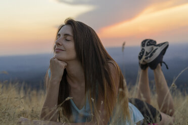 Lächelnde junge Frau mit geschlossenen Augen im Gras liegend bei Sonnenuntergang - AFVF01366