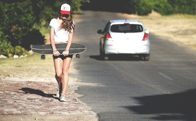 Junge Frau geht mit einem Skateboard am Straßenrand spazieren. - MINF06626