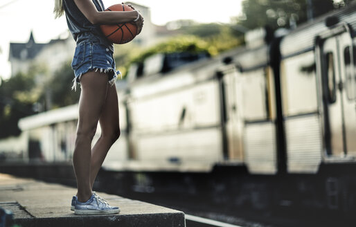 Junge Frau, die auf einem Bahnsteig steht und einen Basketball hält, mit einem Zug im Rücken. - MINF06641