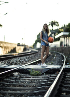 Junge Frau steht auf Eisenbahnschienen und hält einen Basketball. - MINF06642