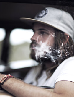 Bärtiger junger Mann mit Baseballmütze, der in einem Auto sitzt und Zigarette raucht, Zigarettenrauch. - MINF06701