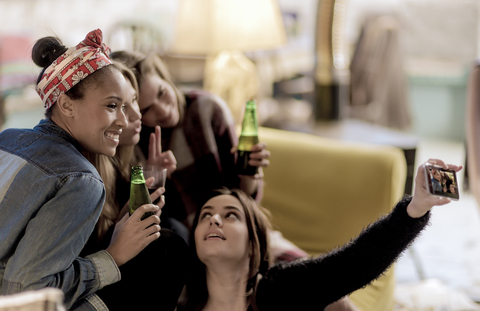 Vier junge Frauen sitzen auf einem Sofa, lachen, machen ein Selfie und halten Bierflaschen in der Hand., lizenzfreies Stockfoto