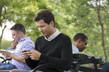 Sommer in der Stadt. Menschen im Freien, die unterwegs in Kontakt bleiben. Eine Frau und drei Männer sitzen im Park, jeder mit seinem eigenen Telefon oder Tablet. - MINF06769