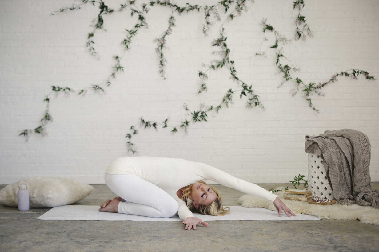 A blonde woman, in a white leotard and leggings, kneeling on a yoga mat  with her