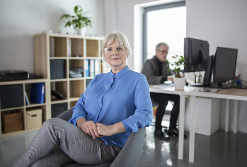 Ältere Geschäftsfrau sitzt in einem Büro mit einem Kollegen, der hinter ihr arbeitet - AWF00178