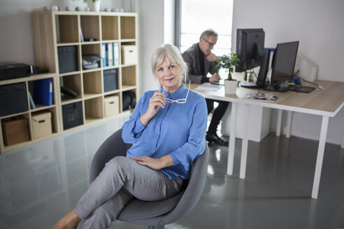 Ältere Geschäftsfrau sitzt in einem Büro mit einem Kollegen, der hinter ihr arbeitet - AWF00179