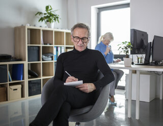 Älterer Mann sitzt im Büro und macht sich Notizen, während ein Kollege hinter ihm arbeitet - AWF00180