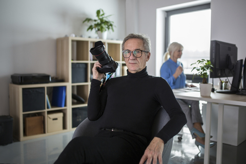 Senior man with camera sitting in office with colleague working behind him stock photo