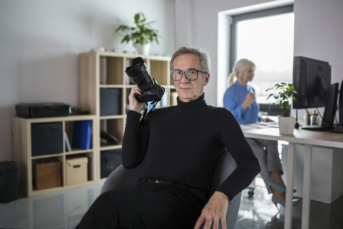 Senior man with camera sitting in office with colleague working behind him - AWF00181