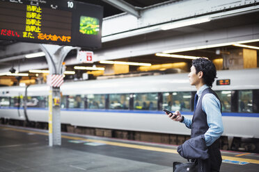 Geschäftsmann in blauem Hemd und Weste steht auf dem Bahnsteig und hält ein Mobiltelefon in der Hand. - MINF06800