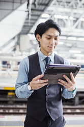 Businessman wearing blue shirt and vest standing on train station platform, holding digital tablet. - MINF06802