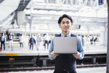 Geschäftsmann in blauem Hemd und Weste steht auf dem Bahnsteig, hält einen Laptop und schaut in die Kamera. - MINF06803