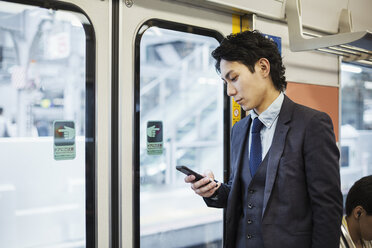 Geschäftsmann im Anzug steht in einem Nahverkehrszug und hält ein Mobiltelefon in der Hand. - MINF06806