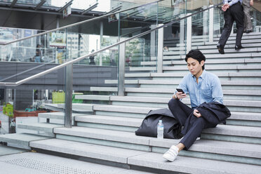 Geschäftsmann in blauem Hemd sitzt im Freien auf einer Treppe und schaut auf sein Mobiltelefon. - MINF06812