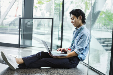 Businessman wearing blue shirt sitting on floor indoors, leaning against glass wall, working on laptop computer. - MINF06818