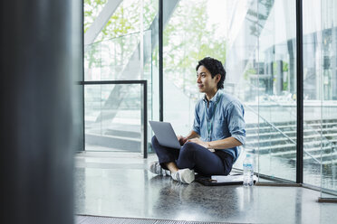 Businessman wearing blue shirt sitting on floor indoors, leaning against glass wall, working on laptop computer. - MINF06820