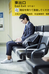 Businessman wearing suit and glasses sitting at train station, working on laptop. - MINF06824