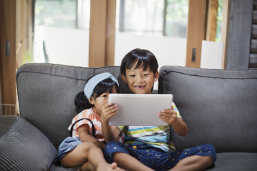 Junge und junges Mädchen mit schwarzen Haaren sitzen auf einem grauen Sofa und schauen auf ein digitales Tablet. - MINF06829