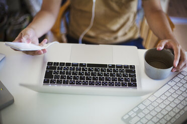 Blick auf eine Person, die mit einem Laptop am Schreibtisch sitzt und ein Mobiltelefon und eine Kaffeetasse hält. - MINF06837