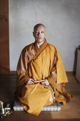 Buddhistischer Mönch mit rasiertem Kopf und goldenem Gewand kniet in einem Tempel auf dem Boden, hält eine Mala und schaut in die Kamera. - MINF06841