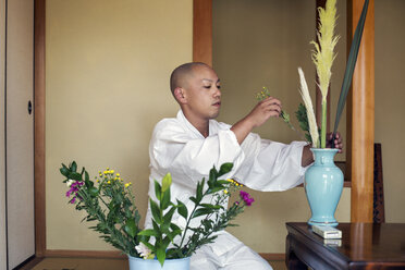Buddhistischer Mönch mit kahlgeschorenem Kopf und weißer Robe kniet auf dem Boden und arrangiert Blumen in einer blauen Vase. - MINF06844