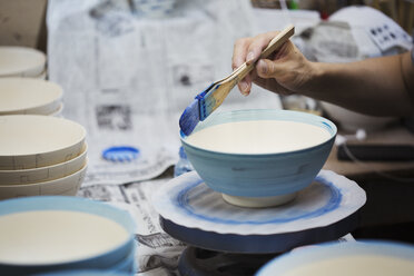 Nahaufnahme einer Person bei der Arbeit in einer japanischen Porzellanwerkstatt, die weiße Schalen mit blauer Glasur bemalt. - MINF06848